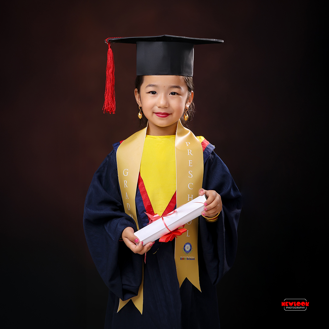 Preschool graduate wearing a black cap and gown with yellow and red sash, holding a rolled-up certificate with a red ribbon, smiling against a dark background